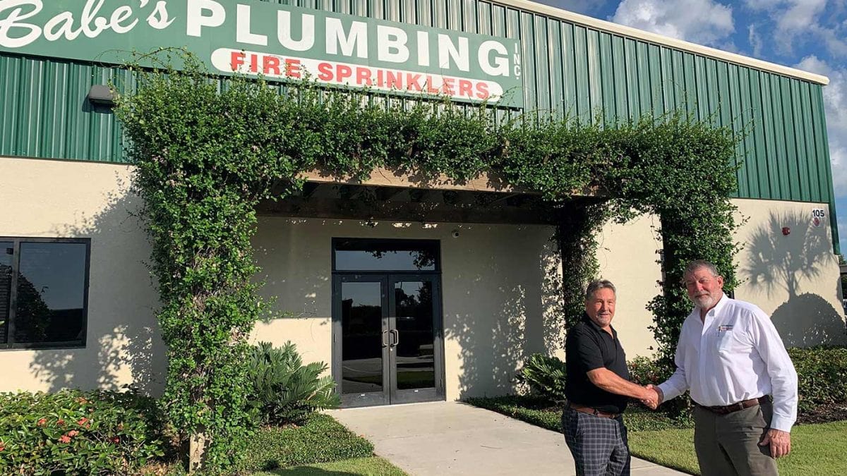 Two men shaking hands in front of a building with signage that reads "Babe’s Plumbing Fire Sprinklers.