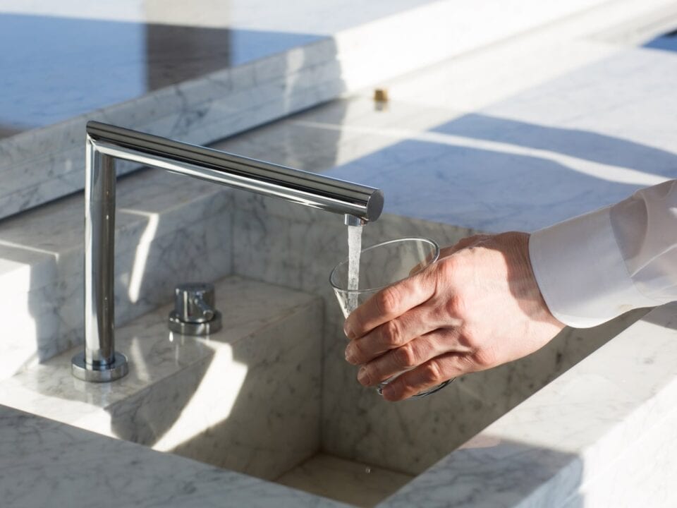 A person in a white shirt filling a glass with water from a kitchen faucet over a marble sink.
