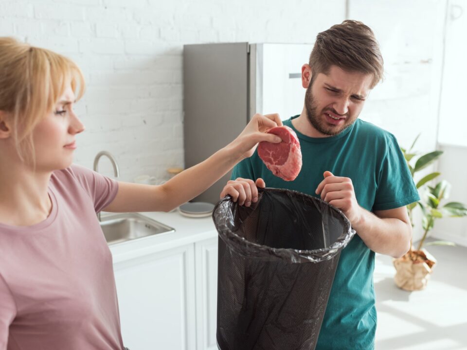 A woman throws food into the garbage disposal as a man holding a trash bin looks at it with a grimace, troubled by the disposal only humming.