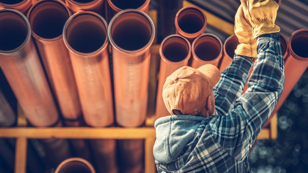 Worker in a hard hat and gloves organizing large polybutylene pipes on a rack.