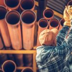 Worker in a hard hat and gloves organizing large polybutylene pipes on a rack.