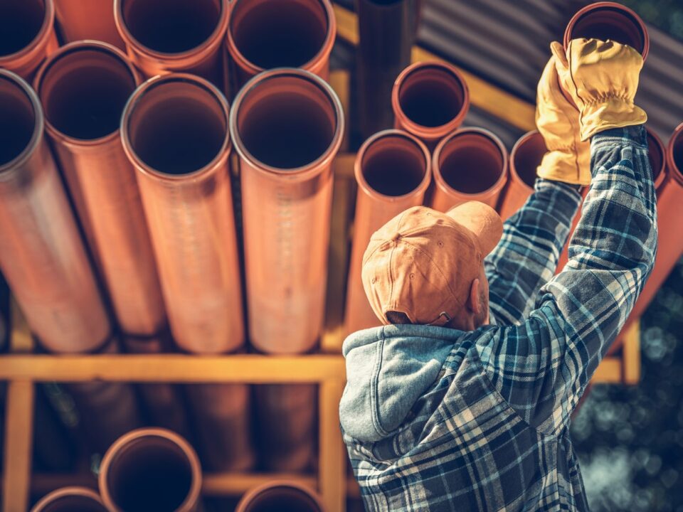 Worker in a hard hat and gloves organizing large polybutylene pipes on a rack.