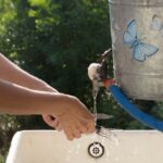 Person washing hands at an outdoor sink, consuming 40 gallons of water from a container with a butterfly sticker.