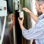 Man collecting a sample from a steel tank at a brewery or factory to monitor clogged water heater drainage.
