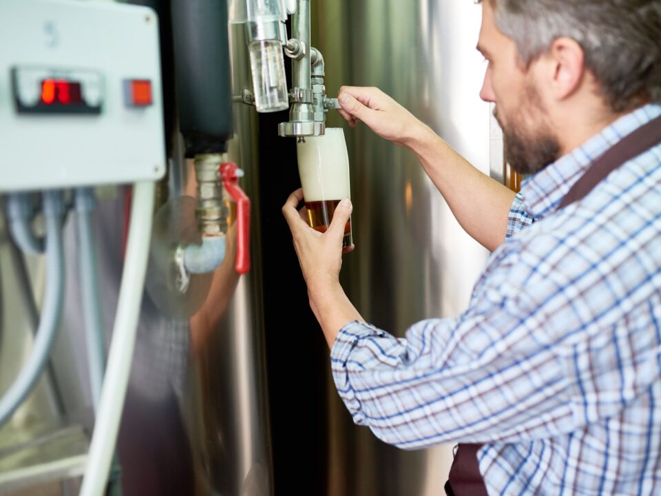 Man collecting a sample from a steel tank at a brewery or factory to monitor clogged water heater drainage.